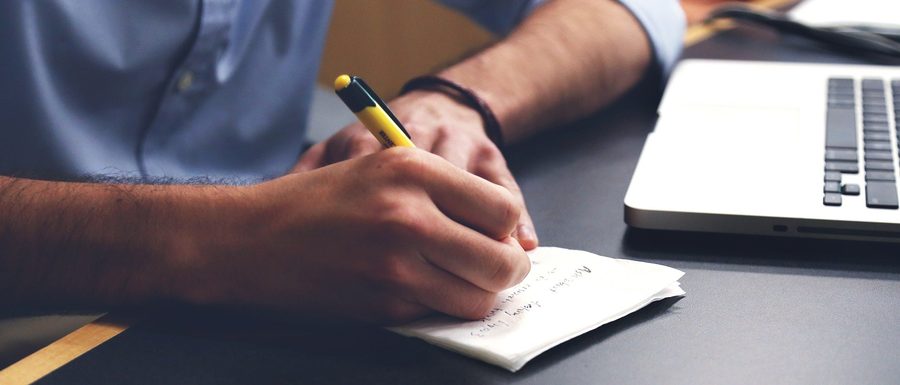 Une main écrivant à la main sur un carnet, près d'un ordinateur